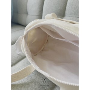 May include: A white canvas tote bag with a zipper closure. The bag is open and the inside is visible. The bag has a white strap.