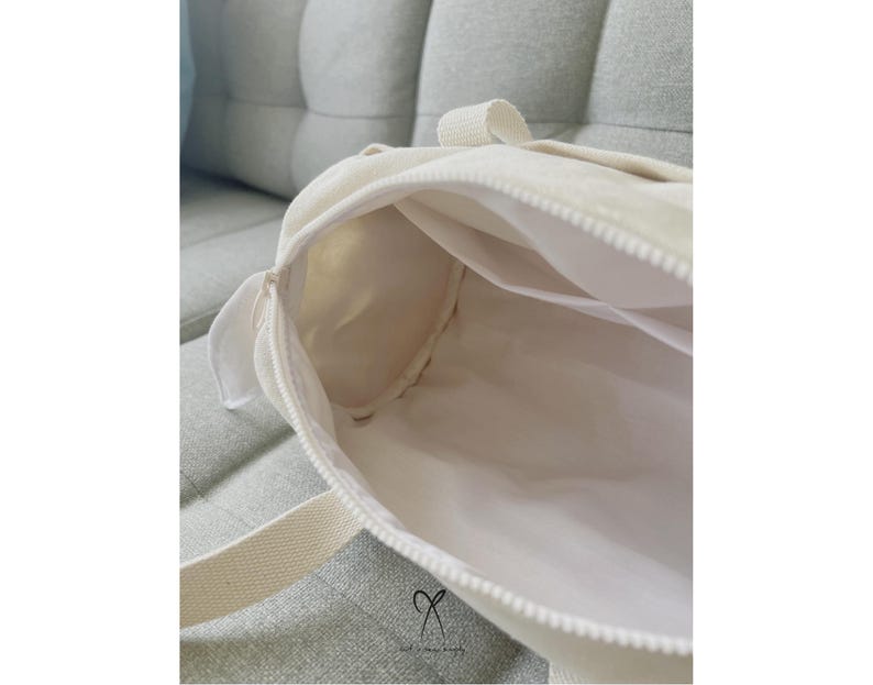 May include: A white canvas tote bag with a zipper closure. The bag is open and the inside is visible. The bag has a white strap.