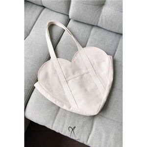 K&ouml;nnte beinhalten: Beigefarbene, herzf&ouml;rmige Tragetasche mit langen Henkeln und einer kleinen Vordertasche. Die Tasche ist aus Canvas gefertigt und liegt auf einem grauen Sofa. Schlichtes, elegantes Design.