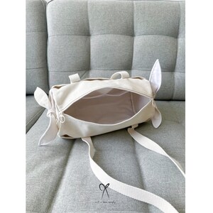 May include: A white canvas tote bag with a zipper closure and two short handles. The bag is open and the inside is visible. The bag is sitting on a light gray couch.
