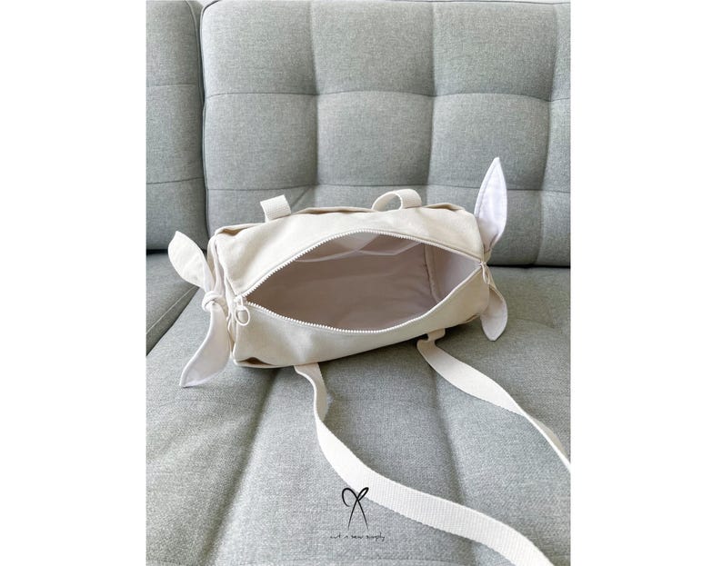 May include: A white canvas tote bag with a zipper closure and two short handles. The bag is open and the inside is visible. The bag is sitting on a light gray couch.
