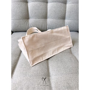 May include: A beige canvas tote bag with a simple design. The bag is sitting on a gray fabric surface.
