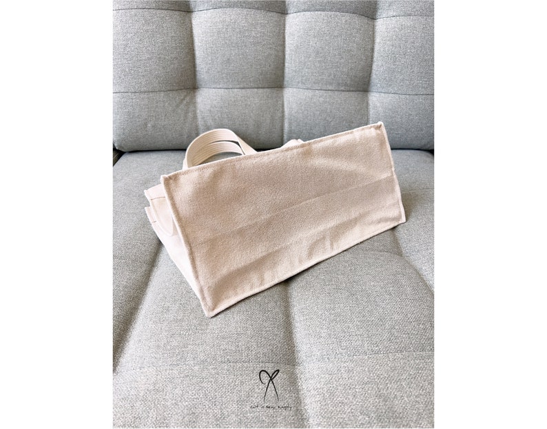 May include: A beige canvas tote bag with a simple design. The bag is sitting on a gray fabric surface.
