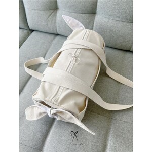 May include: A cream-colored canvas duffel bag with a zipper closure and a bow detail. The bag is sitting on a light gray fabric surface.