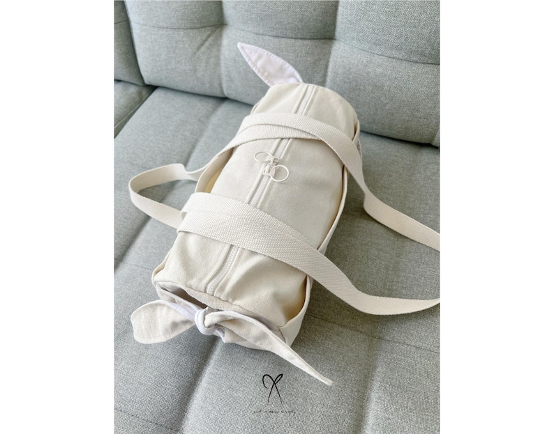 May include: A cream-colored canvas duffel bag with a zipper closure and a bow detail. The bag is sitting on a light gray fabric surface.