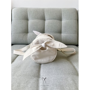 May include: A white canvas tote bag with a knotted strap. The bag is cylindrical in shape and has a simple design.