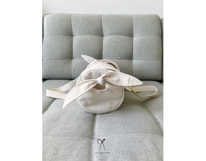 May include: A white canvas tote bag with a knotted strap. The bag is cylindrical in shape and has a simple design.