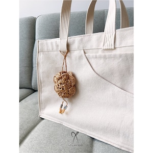 May include: A white canvas tote bag with a brown crocheted flower charm attached to the front. The bag has a large pocket on the front and two handles.