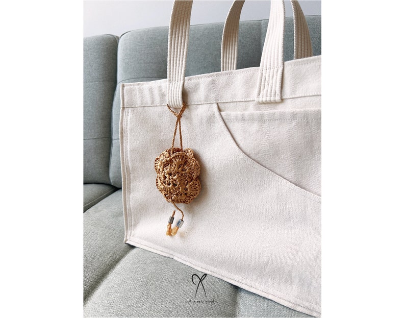 May include: A white canvas tote bag with a brown crocheted flower charm attached to the front. The bag has a large pocket on the front and two handles.