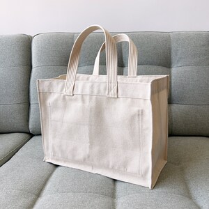 May include: A large, off-white canvas tote bag with thick, white cotton straps. The bag is sitting on a gray couch.