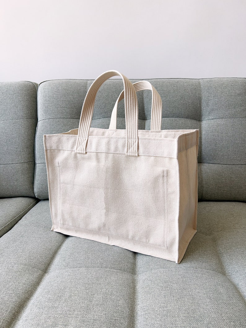 May include: A large, off-white canvas tote bag with thick, white cotton straps. The bag is sitting on a gray couch.