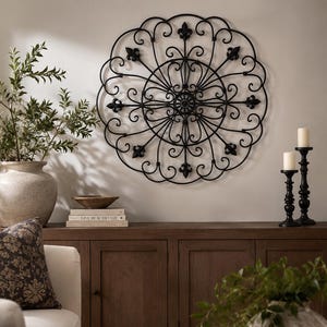 May include: A large, black metal wall art piece with a circular design featuring fleur-de-lis and scrollwork. The artwork hangs above a dark wood cabinet, with decorative vases, candles, and a sofa visible in the room.