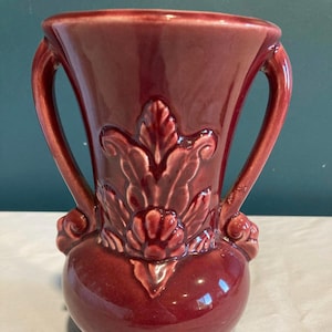 May include: A burgundy ceramic vase with two curved handles. The vase features a raised floral design in the center and a bulbous base. The glossy finish reflects light, highlighting the details of the embossed pattern.