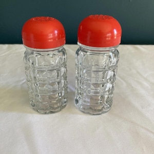 May include: Two clear glass salt and pepper shakers with a textured, grid-like pattern. Each shaker has a bright red, rounded lid. The shakers are displayed on a white surface, suggesting a kitchen or dining setting.
