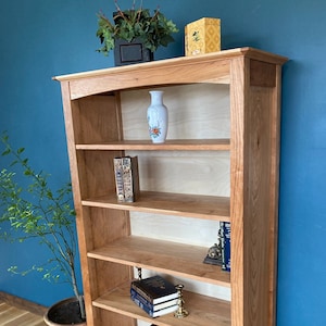 May include: A wooden bookcase with five shelves, featuring a decorative vase with floral designs, books, and a small wooden box. The bookcase is made of cherry wood and has a natural finish. The bookcase is standing on a wooden floor.