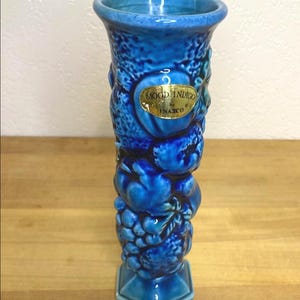 May include: A tall, blue ceramic vase with a textured surface and a hexagonal base. The vase is adorned with relief fruit designs and a gold label that reads "MOOD INDIGO by INARCO".
