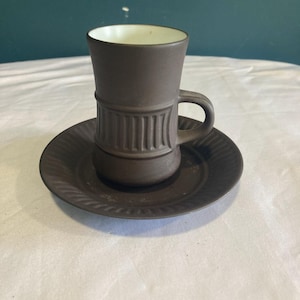 May include: A brown ceramic coffee cup and saucer set with a white interior. The cup has a fluted design around the middle and a handle. The saucer has a fluted edge.