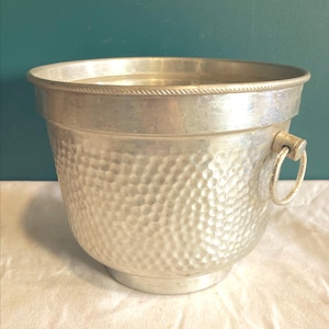May include: A silver metal ice bucket with a hammered texture. The bucket has a rounded shape with a flared rim and a small handle. The bucket is sitting on a white surface.