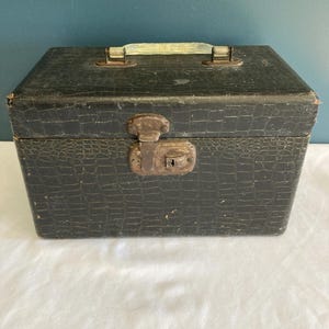 May include: A vintage black box with a faux crocodile skin texture. The box has a metal clasp and handle. The handle is a light green color. The box is sitting on a white surface.