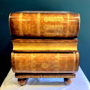 May include: A unique, two-tiered side table designed to resemble a stack of antique books. The table features a warm brown finish with gold accents and decorative text, resting on small, rounded feet. The top tier reads "NEW YORK".