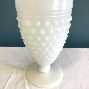 Puede incluir: Una copa de vidrio blanco con diseño hobnail, con un borde ancho y un cuerpo texturizado en forma de cono. La copa tiene una base redonda y un tallo corto. El vidrio tiene un acabado brillante. Se encuentra sobre una superficie blanca y un fondo azul oscuro.