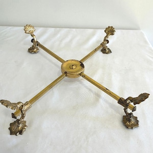 May include: A gold-toned metal cross-shaped stand featuring four adjustable arms extending from a central circular hub. Each arm terminates in a decorative claw-style foot with a flat, scalloped base, providing stability for the structure.