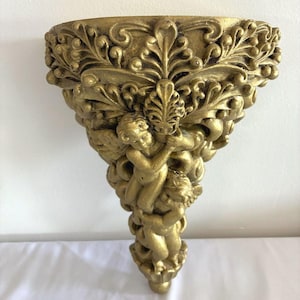May include: A gold-colored wall shelf featuring intricate relief carvings of two cherubs surrounded by ornate acanthus leaf and scrollwork patterns. The semi-circular top provides a flat surface, while the base tapers into a decorative point.