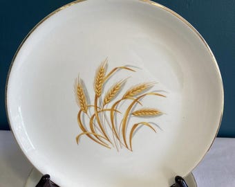 May include: White ceramic plates with a gold rim and a wheat stalk design in shades of gold and brown. The plates are displayed on a dark brown plate stand against a dark teal background.