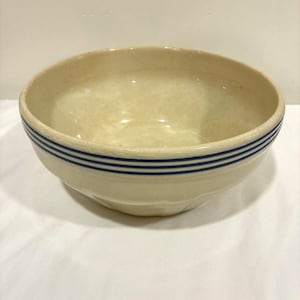 May include: Cream-colored ceramic bowl with three horizontal blue stripes around the rim. The bowl has a rounded shape and a slightly textured surface. A classic kitchenware item.