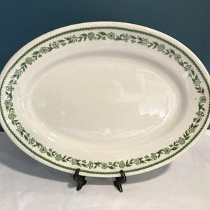 May include: Oval, white serving platter with a green floral border. The platter is supported by a black metal stand. The green border features a repeating pattern of flowers and leaves.
