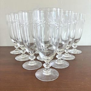 May include: A set of clear, stemmed champagne flutes. Each glass features an etched floral design and a round base. The glasses are arranged in a group on a brown surface, showcasing their elegant shape and delicate details. Ideal for special occasions.