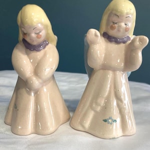 May include: Two ceramic angel figurines. Each angel has blonde hair, closed eyes, and a purple collar. One angel has hands clasped, the other has arms outstretched. Both wear light pink robes. A small blue flower is painted on one robe.