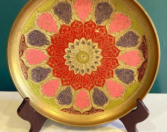 May include: A round, gold-colored decorative tray with an intricate floral design. The design features a central gold medallion surrounded by red, pink, purple, and green petal-shaped elements. The tray is displayed on a brown wooden stand.