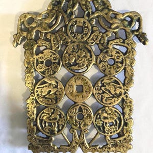 May include: An antique brass-colored decorative plaque featuring nine circular coin-like elements with intricate designs. The plaque has an ornate, openwork border with stylized animal and floral motifs. The overall design suggests a vintage or Asian-inspired aesthetic.