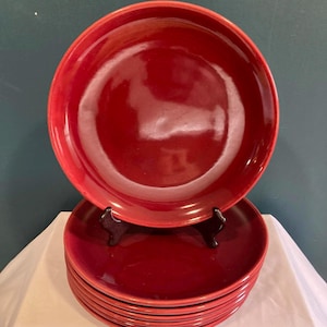 May include: A stack of red ceramic plates. The plates are a deep, glossy red color. One plate is elevated on a black stand, showcasing the design. The plates are arranged on a white surface against a dark green background.