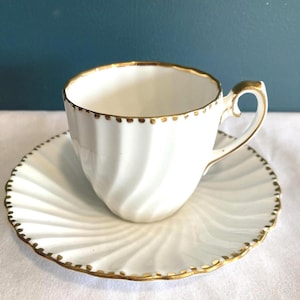 May include: White teacup and saucer set with gold trim. The teacup has a spiral design and a gold handle. The saucer also has a spiral design and gold trim. The set is on a white surface.