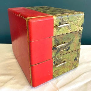 May include: A small, three-drawer box with a red front and a green and gold patterned top and drawer fronts. The pattern features cranes. The drawers have small, silver-colored handles. The box is rectangular and appears to be made of wood.