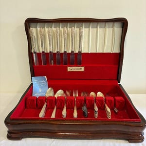 May include: A wooden silverware chest with a red velvet interior containing silver-plated flatware. The lid features a white pleated lining and a Community brand plaque. A blue guarantee card is tucked inside the case with the utensils.