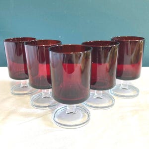 May include: Six ruby red wine glasses with clear stems and bases are arranged on a white surface. The glasses have a cylindrical shape and are evenly spaced. The background is a teal color.