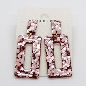 May include: A pair of rose gold rectangular earrings with a glitter finish. The earrings have a small, open rectangle shape in the center.