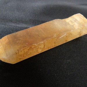 Puede incluir: Un cristal largo, translúcido, marrón claro con un extremo puntiagudo. El cristal tiene una superficie rugosa y texturizada.