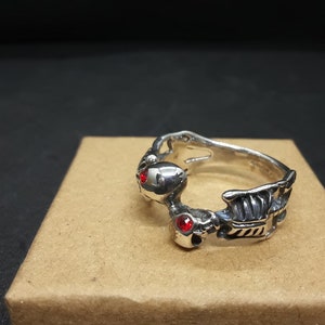 May include: A silver-colored ring with a unique design. The ring features two small, stylized skulls with red gemstone eyes. The band has an intricate, textured pattern. The ring is displayed on a brown surface against a dark background.