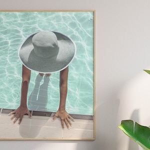 Puede incluir: Una mujer con un gran sombrero de sol gris de ala ancha emerge de una piscina turquesa. Sus manos están en el borde de la piscina y su sombra es visible en el agua.