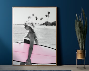 May include: A black and white photo of a woman holding a pink surfboard on a beach with palm trees in the background.