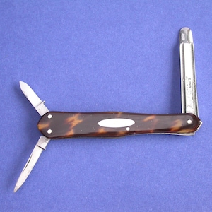 May include: A vintage pocket knife with a tortoiseshell handle and two blades extended. A silver ruler is also extended. The knife has a brown and cream pattern on the handle and is set against a blue background.