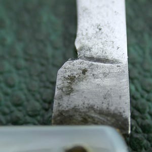 以下が含まれることがあります： ヴィンテージのストレートカミソリの刃のクローズアップ。金属製の刃は、欠けたエッジと鈍いマット仕上げを示しています。背景は、質感のある濃い緑色の表面です。