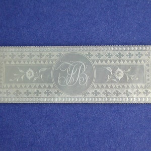 May include: A rectangular, white, mother-of-pearl bookmark with an intricate, engraved design. The design features a floral motif, a circle with the initials "FB" inside, and a border of small circles and triangles.