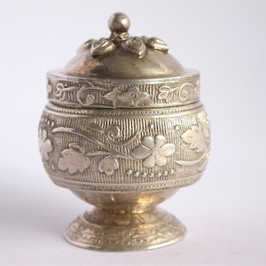 以下が含まれることがあります： 花柄の装飾が施された銀製の蓋付きの壺。壺は丸い胴体とドーム型の蓋を持ち、蓋の上にはつまみがある。