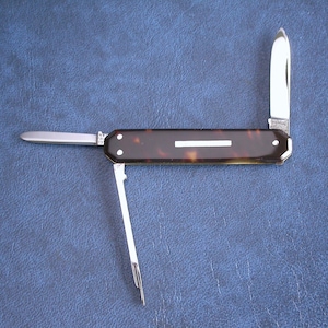 May include: A pocket knife with a tortoiseshell handle and three open blades. The silver blades contrast with the dark handle, which features a white stripe. The knife is displayed on a blue surface.