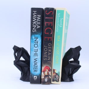 May include: Black geometric bookends holding three books. The book titles are "Into the Water" by Paula Hawkins, "Siege" by Geraint Jones, and "Sea of Lost Love" by Santa Montefiore.